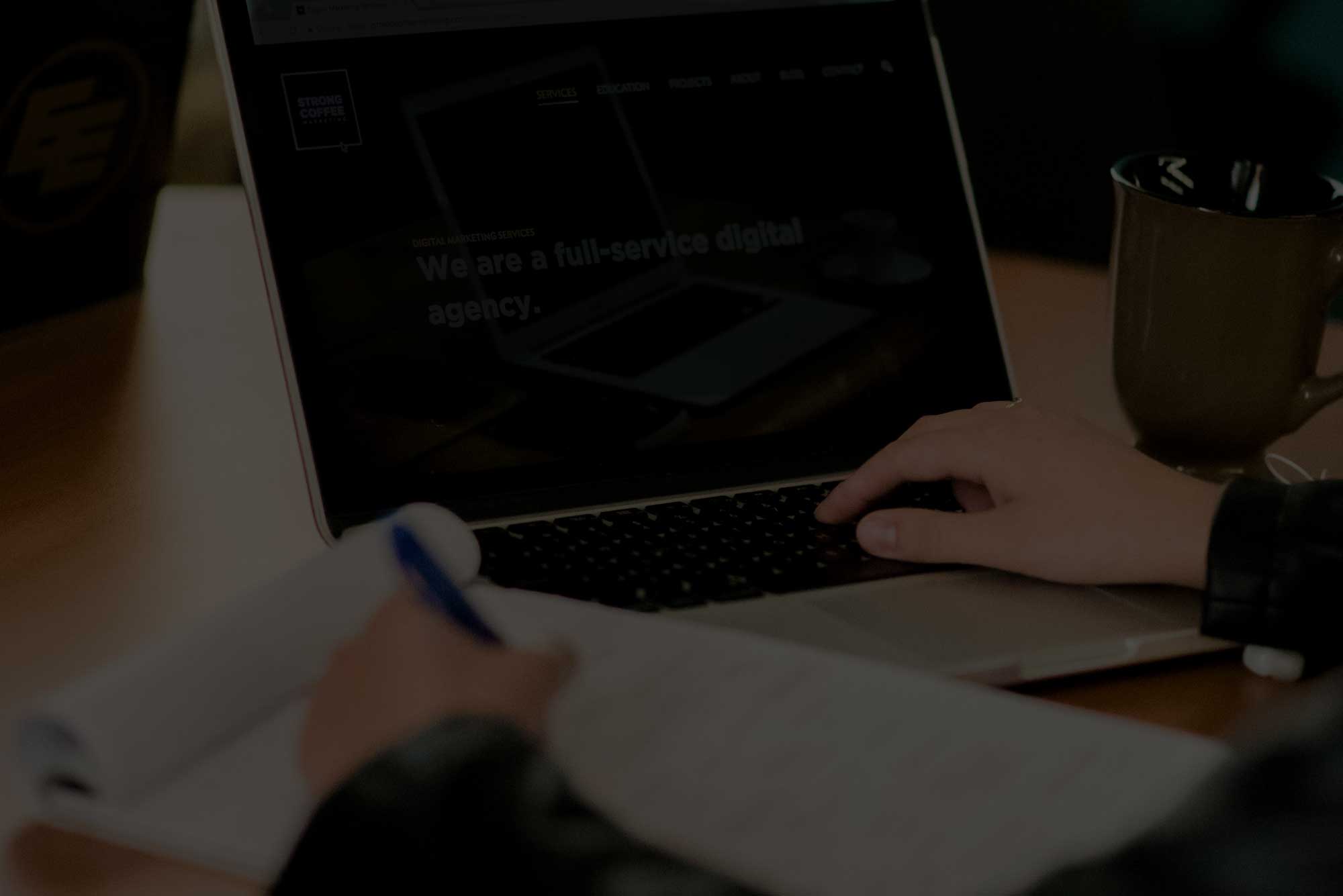 Close-up of a person type on a laptop while taking notes on paper with the other hand.