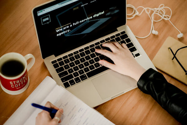 Overhead view of a person working on a laptop and taking notes. A Strong Coffee mug (with coffee) is visible in the frame.